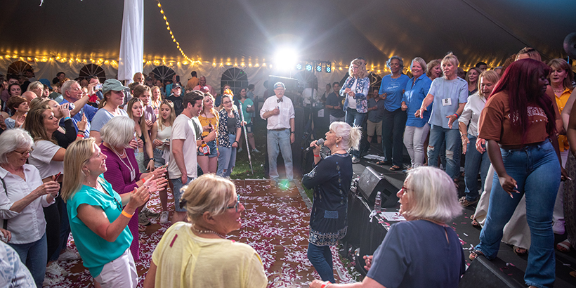 Woman holds microphone as she is surrounded by people on the stage and dance floor