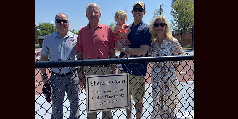 Four people stand on a tennis court, one holding a child