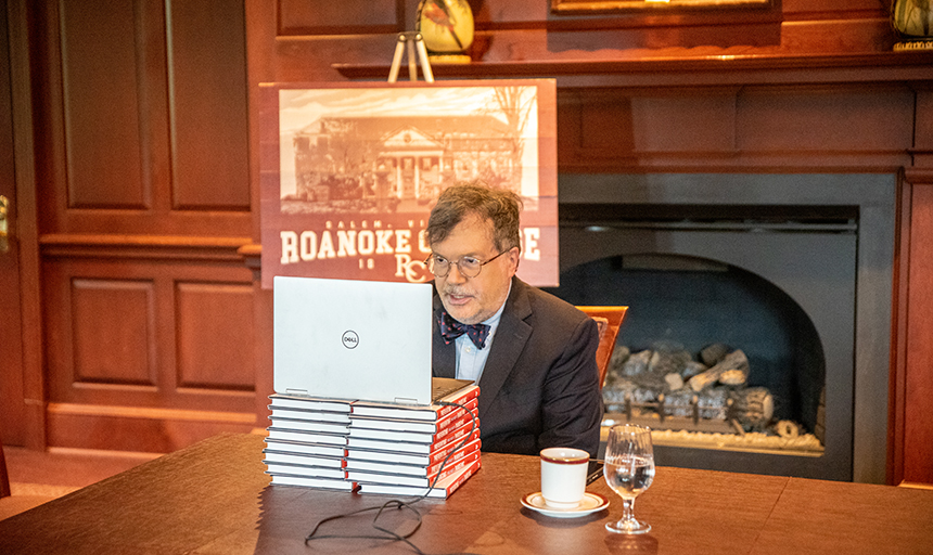 Man in suit sits at table with computer in front of him and Roanoke College sign behind him