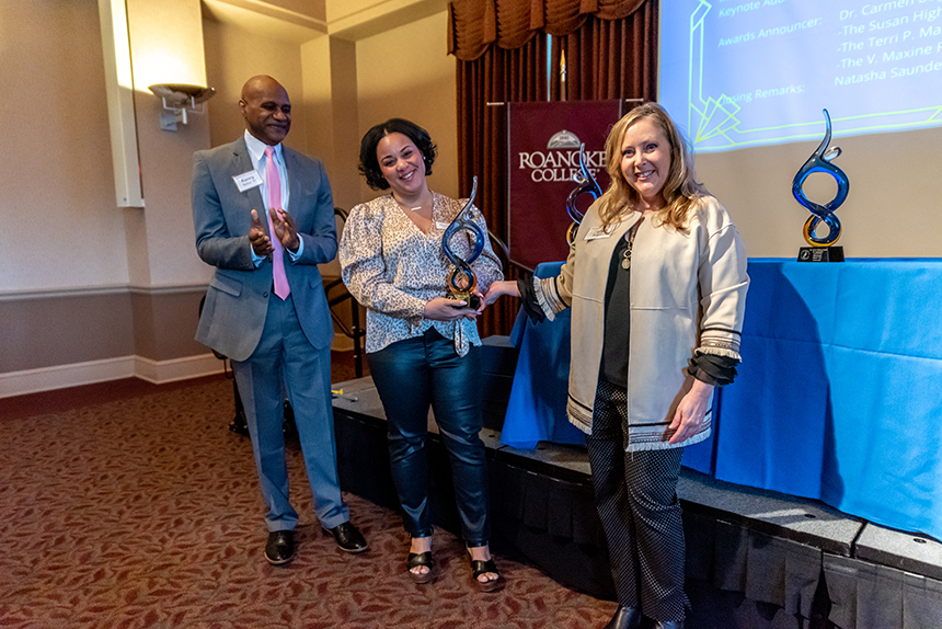 two women and a man pose with award in front of Roanoke College banner