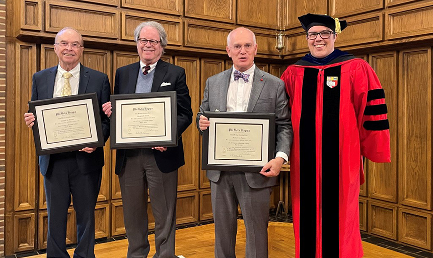 Four men stand together, three of them holding PBK certificates in frames. Fourth man is wearing academic cap and gown