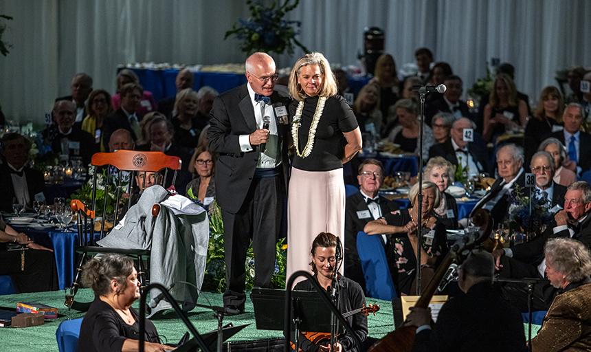 Mike and Terri Maxey stand on a stage, he is holding microphone, and they are surrounded by people seated at a dinner