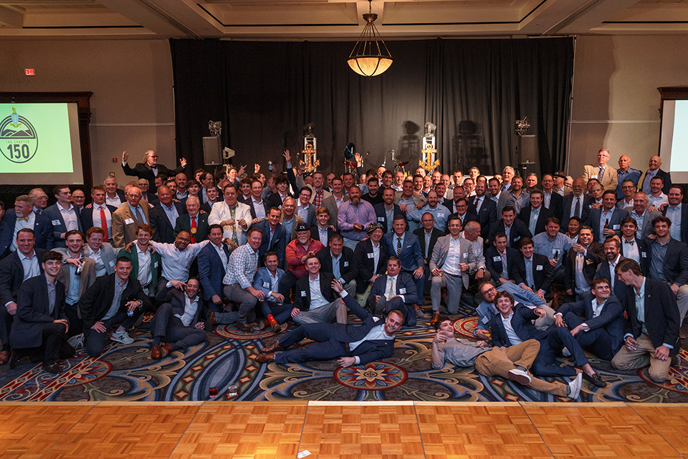 A few dozen men in suits pose for photo in ballroom in front of sign that says "150"