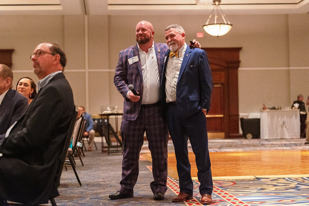 Two men in suits stand smiling together watching something on stage