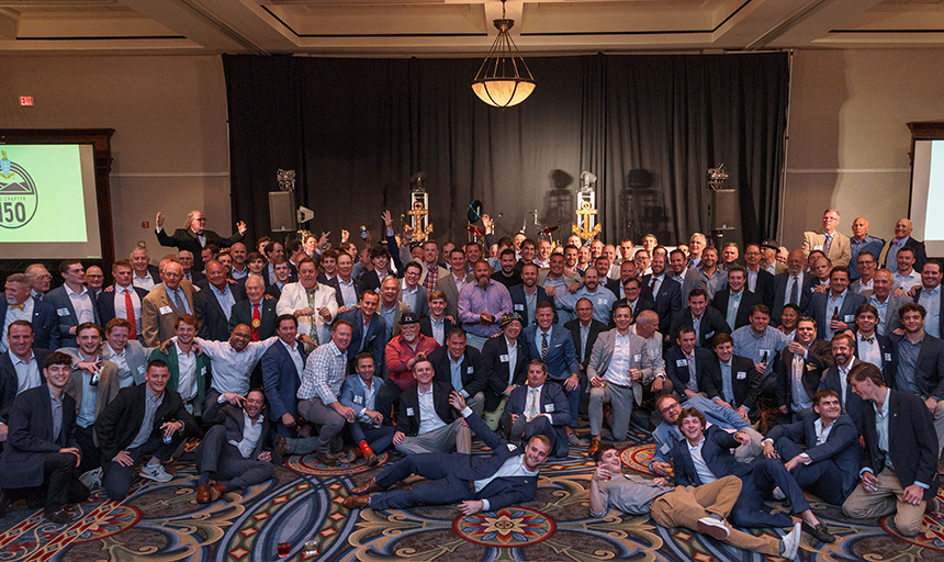 A few dozen men in suits and ties pose for picture in ballroom in front of sign that says "150"