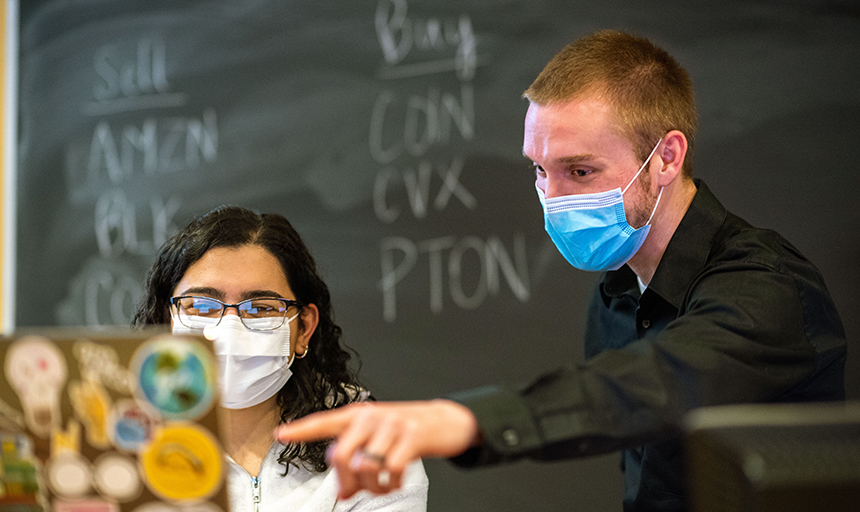 Professor points to laptop with "buy" and "sell" written chalkboard behind him