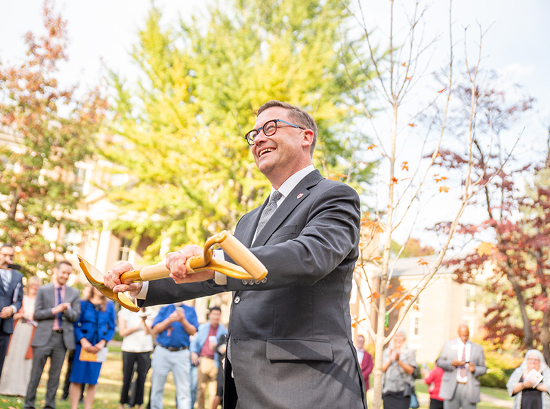 president shushok smiles while holding a gold shovel as the tree planting ceremony begins