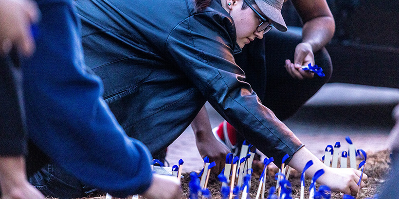 A student leans forward to plant a stake in the ground bearing the name of an enslaved person and adorned with a blue ribbon