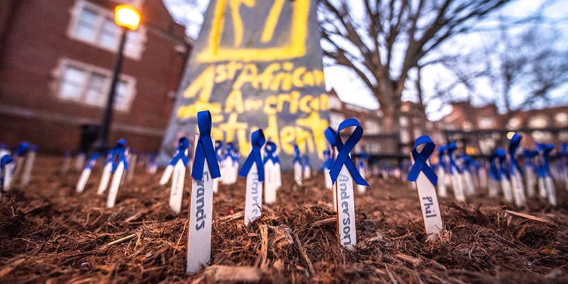 Small stakes bearing the names of enslaved people and adorned with blue ribbons dot the ground