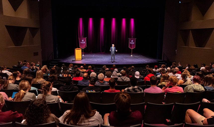 Photo shot from the back of an auditorium full of people, with a man on stage under the lights. 