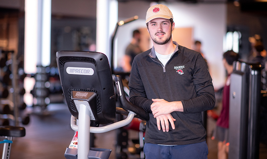 Young man in a black pullover and tan hat leans against an exercise bile and smiles at the camera.