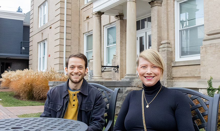 Male student in black jacket sits at an outdoor table next to a female professor in a black sweater, and they are both smiling at the camera. A tan brick building is in the background.