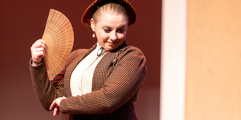 An actor smiles while cooling herself with a small handheld fan during a scene