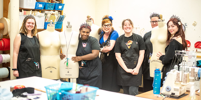 Members of the wardrobe crew, many wearing tiaras, pose next to dressing mannequins backstage