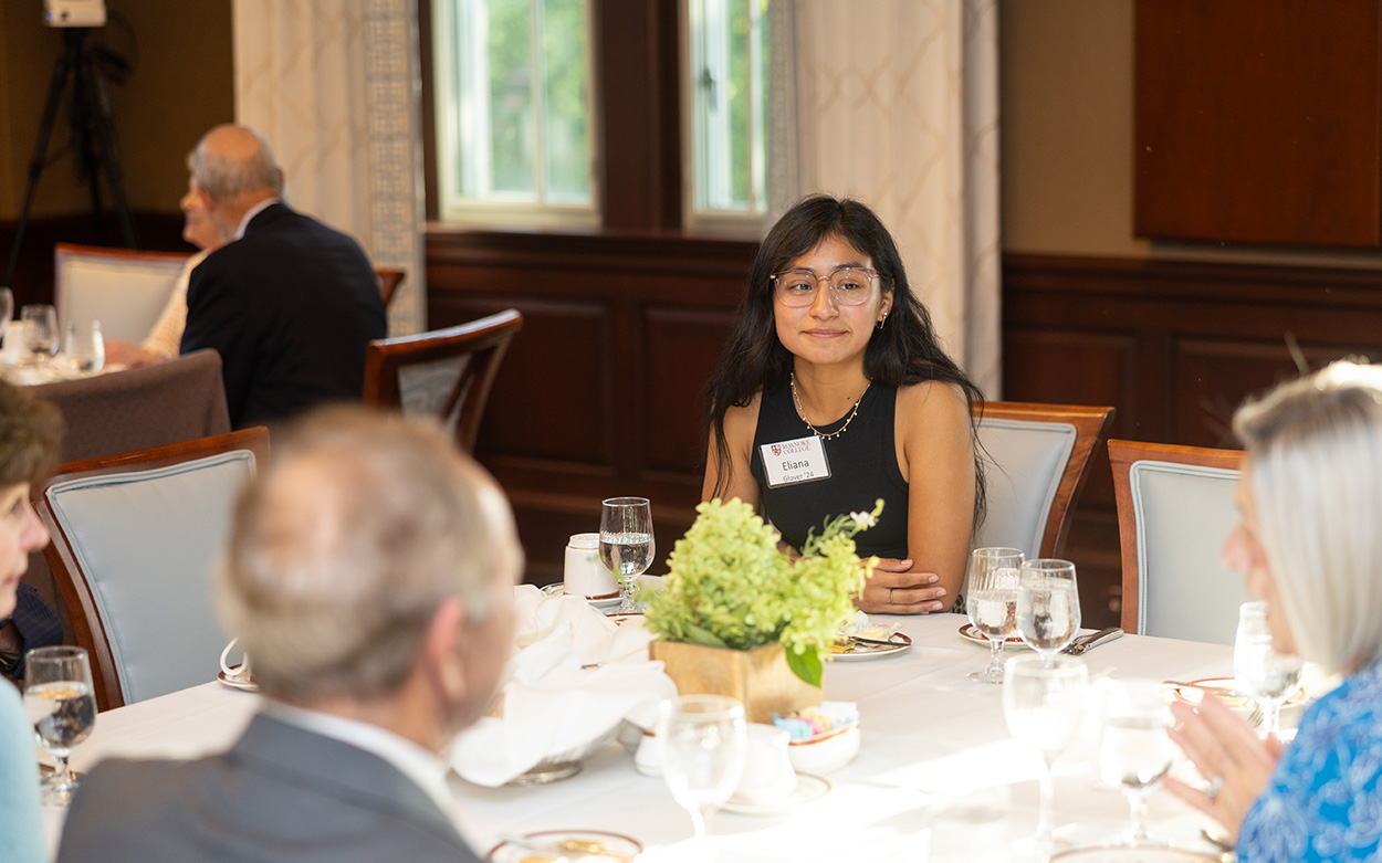 A student smiles while seated with community members around a dining table