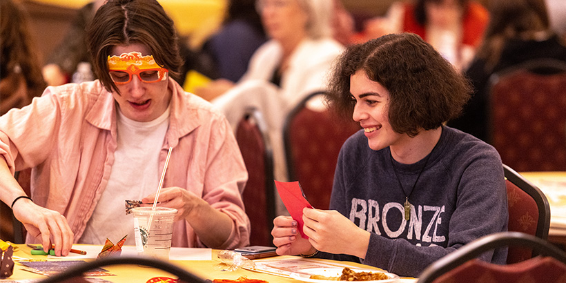 Two students, one wearing a festive set of plastic sunglasses commemorating Lunar New Year, work with pieces of paper to create origami figures