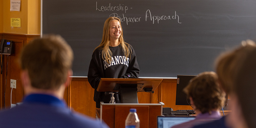 A student smiles while talking at the head of the class in front of a chalkboard bearing the words: Leadership-Behavior Approach