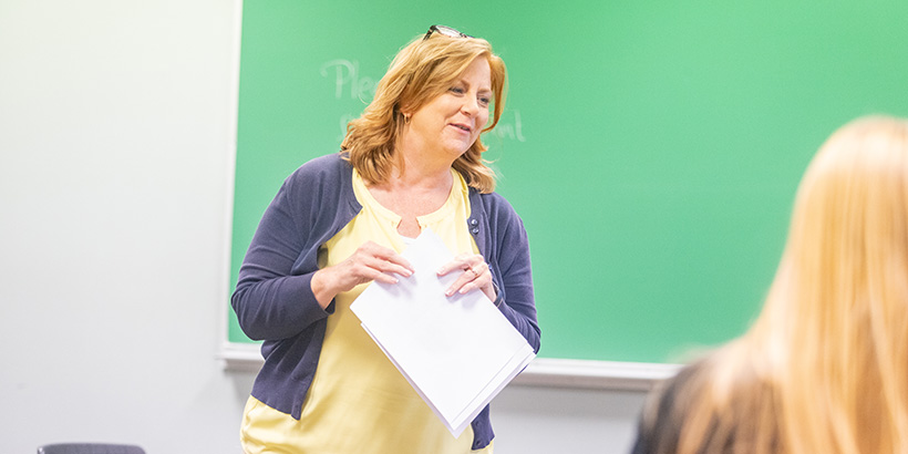 A professor holding sheets of paper smiles while addressing the class