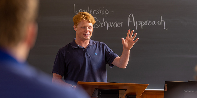 A student gestures while talking from a podium at the front of the classroom