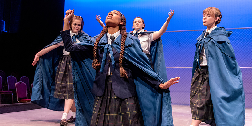 Actors dressed in school uniforms and vivid cloaks raise their arms skyward on stage