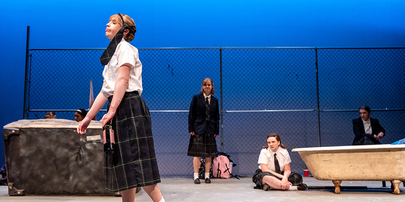 An actor with a necktie wrapped around her head and a bottle of alcohol in her hand lumbers drunkenly across the stage as other cast members look on