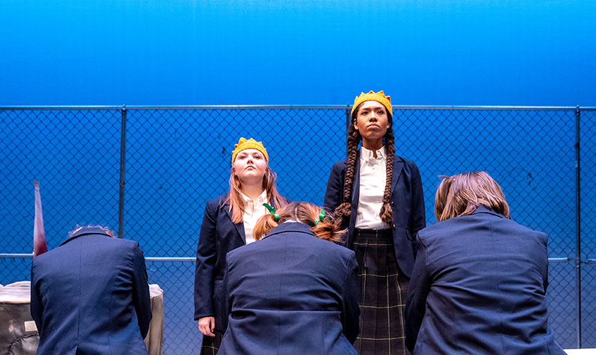 Two actors wearing crowns stand regally while others kneel before them