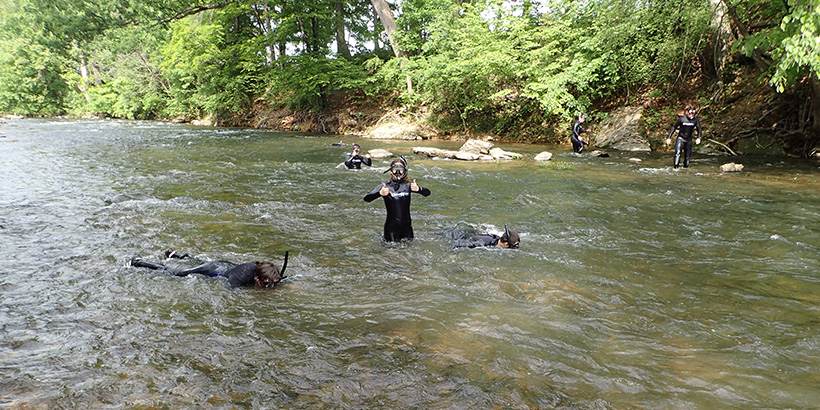 A student wearing snorkeling gear stands in the middle of a rushing river and gives a thumbs up sign with her hands