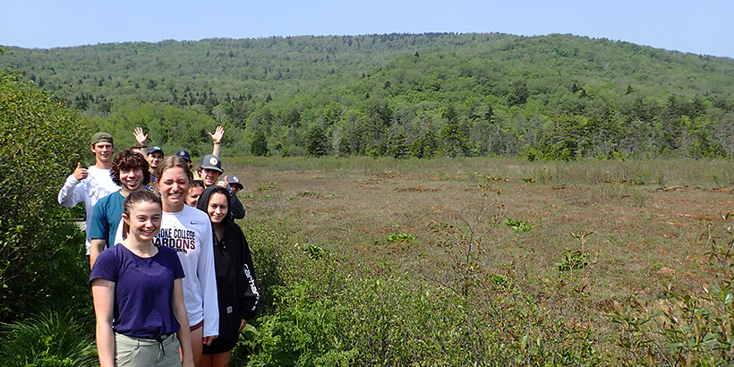 Students smile and wave for a photo during a stop along a trail bordered by a wide open field and a mountain view