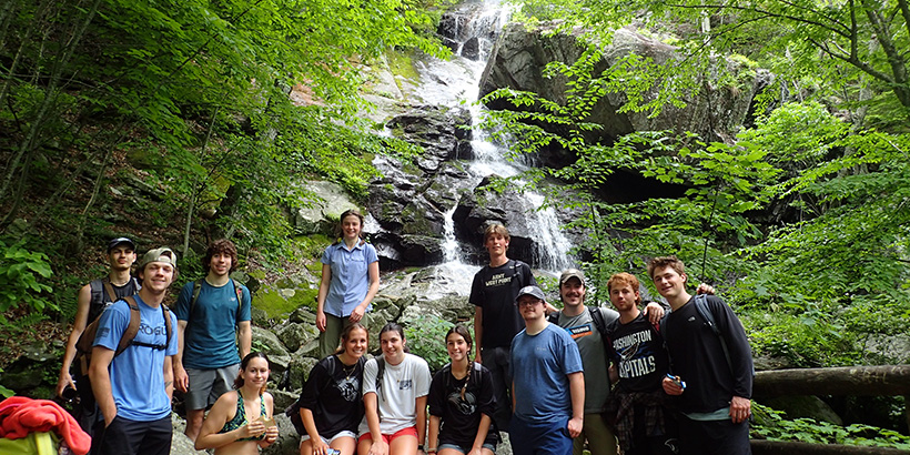 Students pause for a group photo as a waterfall cascades down a craggy rock wall behind them