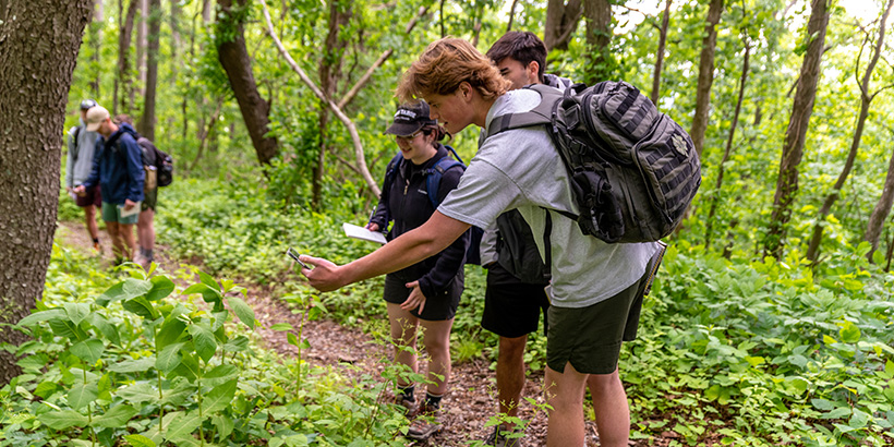 Three students, one leaning down with his hand outstretched, inspect a plant along a mountain trail