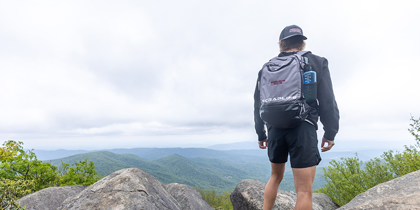 A student carrying a backpack stands near the edge of a mountain overlook with a view of a lush valley below