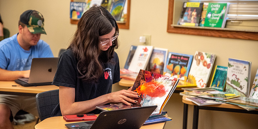 A student pages through a colorful book with other materials and a laptop spread out in front of her on her desk