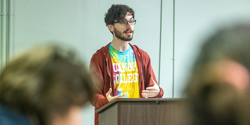 A student in a tie-dyed t-shirt bearing the words Roanoke College addresses the class from a podium during a debate