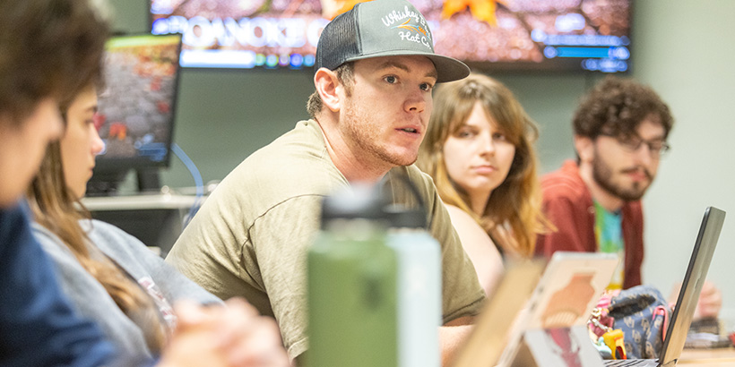 Students sitting at a long table turn to look at one student who is making a point during a debate