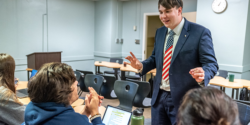 A student wearing a suit and a U.S. flag tie gestures while talking with another student