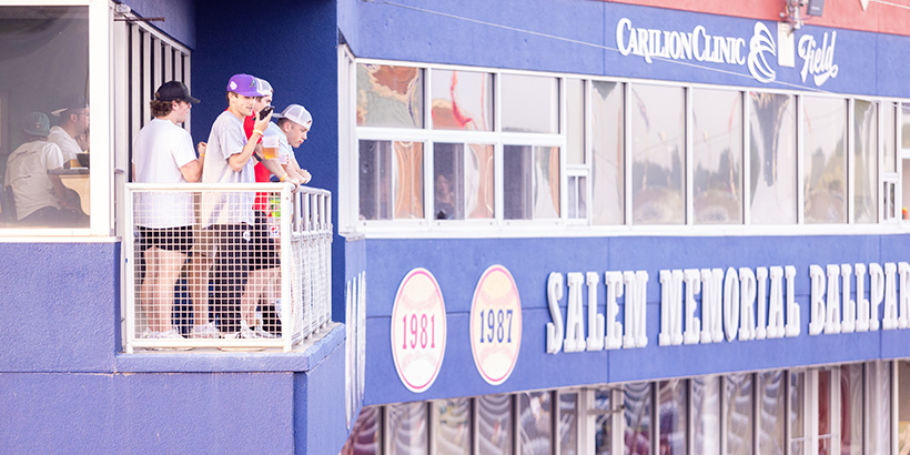 Students lean against the railing of the outdoor seating at a stadium suite of the Salem Red Sox