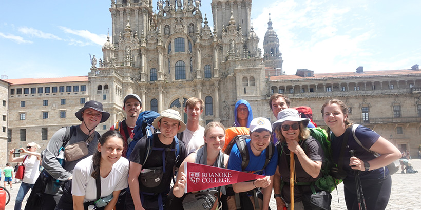 Students holding a Roanoke College pennant flag smile for a photo outside a historic stone building