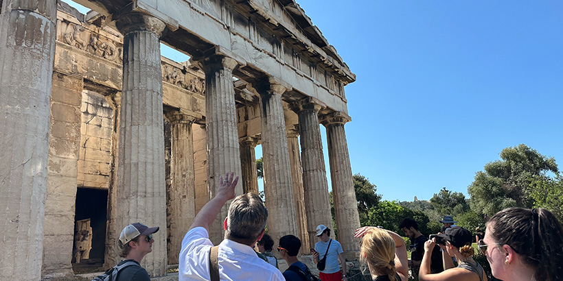 A professor speaks to students while gesturing toward a historical structure lined by Greek columns