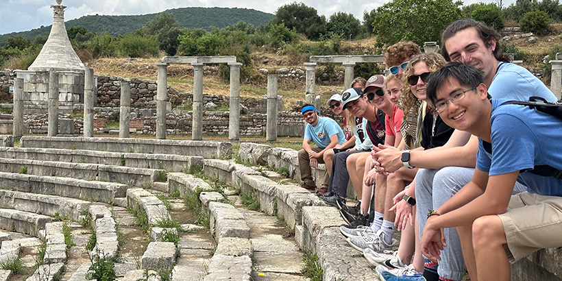 Students sitting in an ancient stone amphiteater smile for a photo