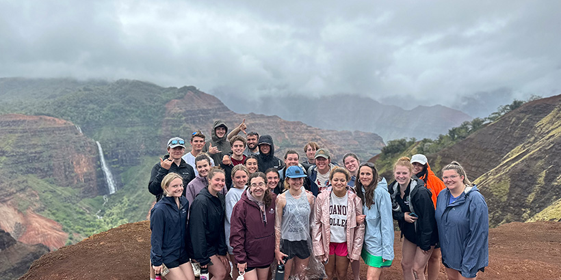 Students smile for a group photo at an overlook with a view of a tropical landscape and a waterfall in the distance