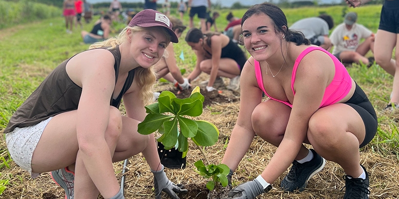 Two students smile while placing a tall plant into a freshly dug foliage bed