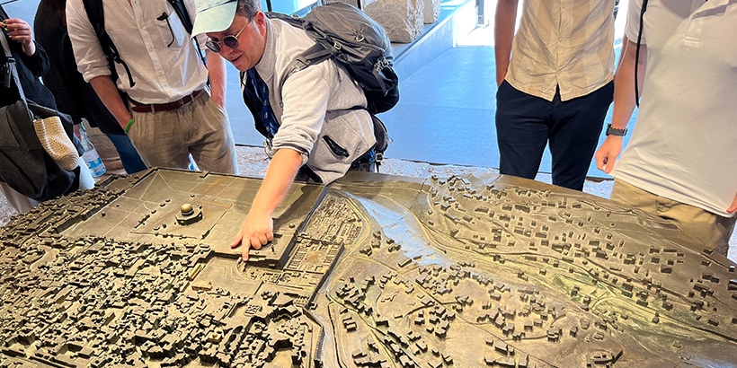 A professor speaks to students while gesturing at an exhibit of a historic city