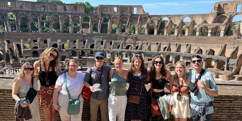 Students smile for a photo with an ancient coliseum-like structure in the background