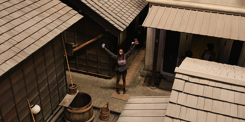 A student standing along a narrow path weaving among wooden buildings smiles and raises her arms skyward