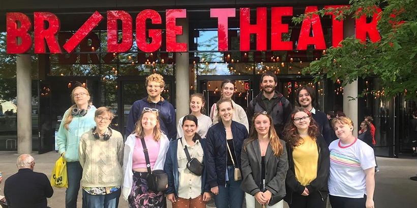 Students smile for a group photo outside the Bridge Theatre, whose name is spelled in large, brightly colored letters in the background
