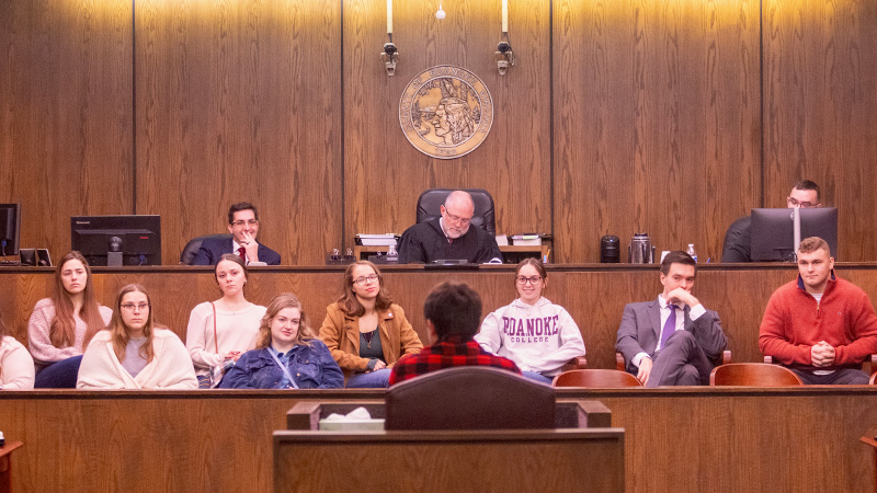 Students fill the seats of a courtroom jury box and listen as a mock trial team member testifies from the witness stand. Judge Geddes, dressed in judicial robes, presides from his bench next to the jury box.