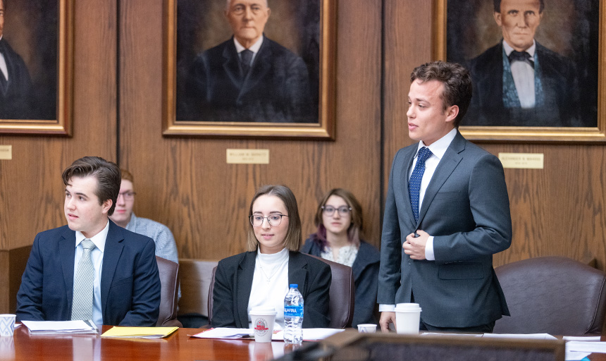 Three students dressed in suits occupy the defense table at a mock trial exhibition held in a wood-paneled courtroom