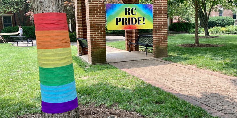 Foreground: A tree on the quad is wrapped in rainbow-hued streamers. Background: A banner that reads RC Pride! hangs from the quad bell tower.