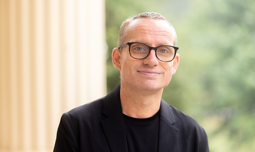 Robert Weiss, wearing a black shirt and blazer with black-framed glasses, stands in front of a column for a portrait.