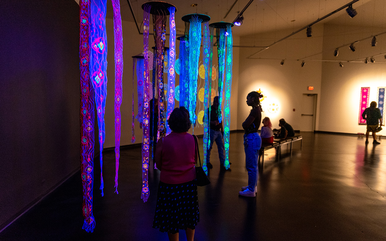 Visitors gaze at an exhibit of embroidered hands dangling from the ceiling against a backdrop of neon light